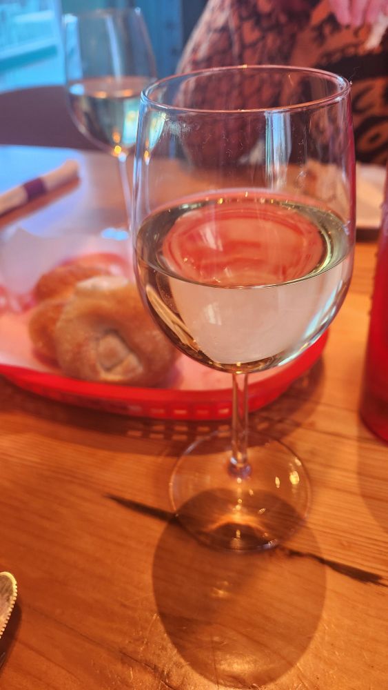 Picture of wine glasses on a wooden table filled with white wine. In the middle is a red basket holding twisted rolls.