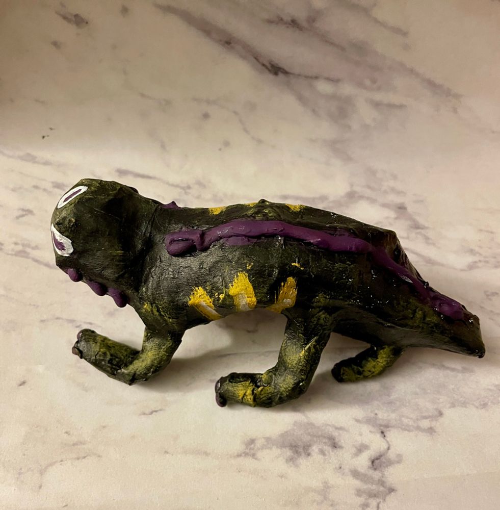 A black painted lizard looking creature. It has purple spines and yellow stripes. Its eyes are like a goat’s, with horizontal purple pupils. It has three sets of legs and a short tail. It’s facing perpendicular to the camera.
