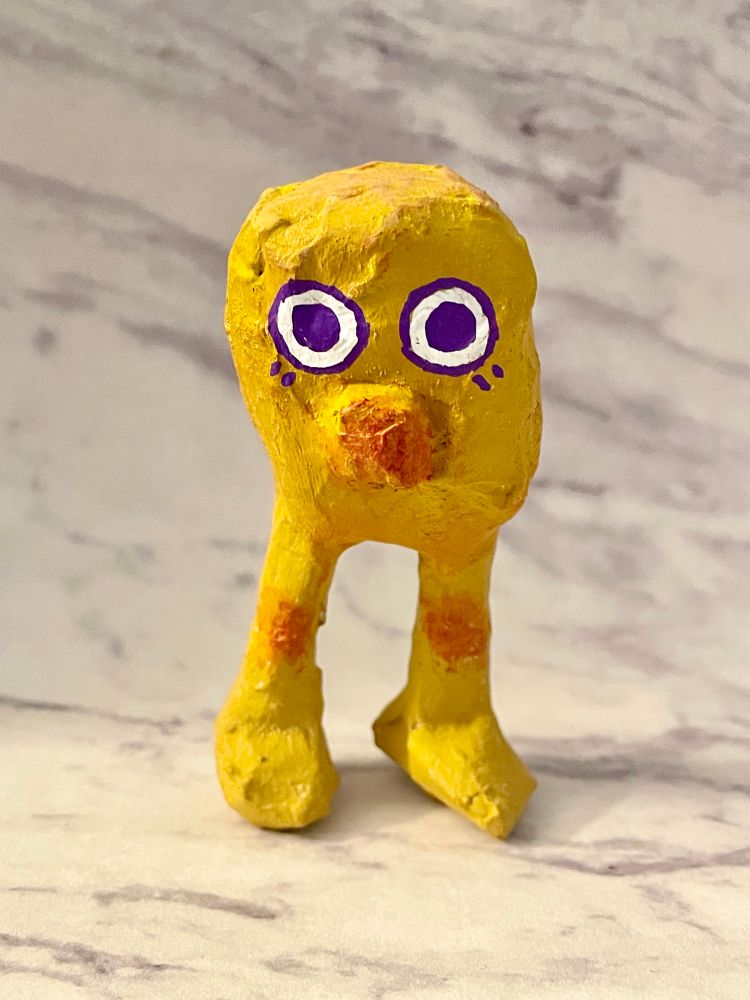 A papier maché creature that looks sort of like a chicken nugget with legs. It has a rounded body and is painted yellow, with a bulbous, orange-tinted nose and big purple eyes. Its knees are also tinted orange. This creature has no arms.