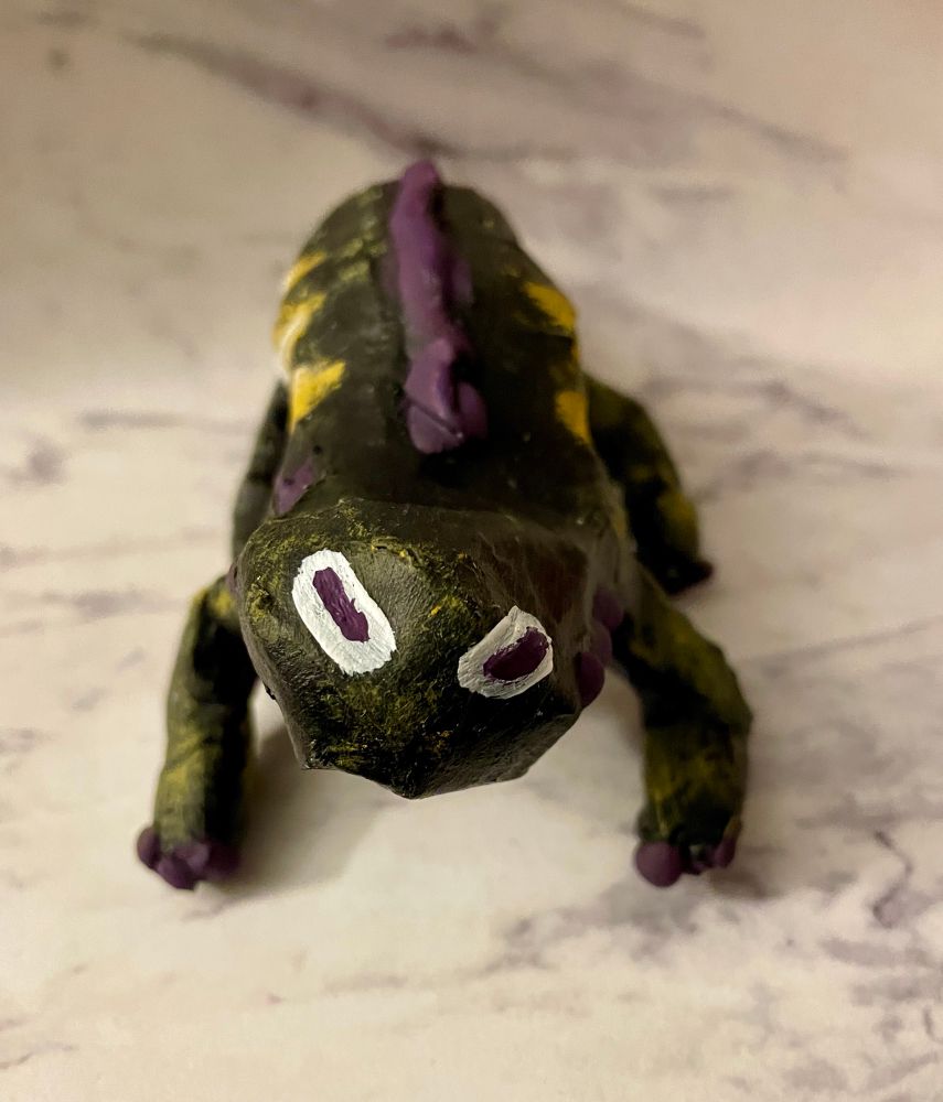 A black painted lizard looking creature. It has purple spines and yellow stripes. Its eyes are like a goat’s, with horizontal purple pupils. It’s facing toward the camera.