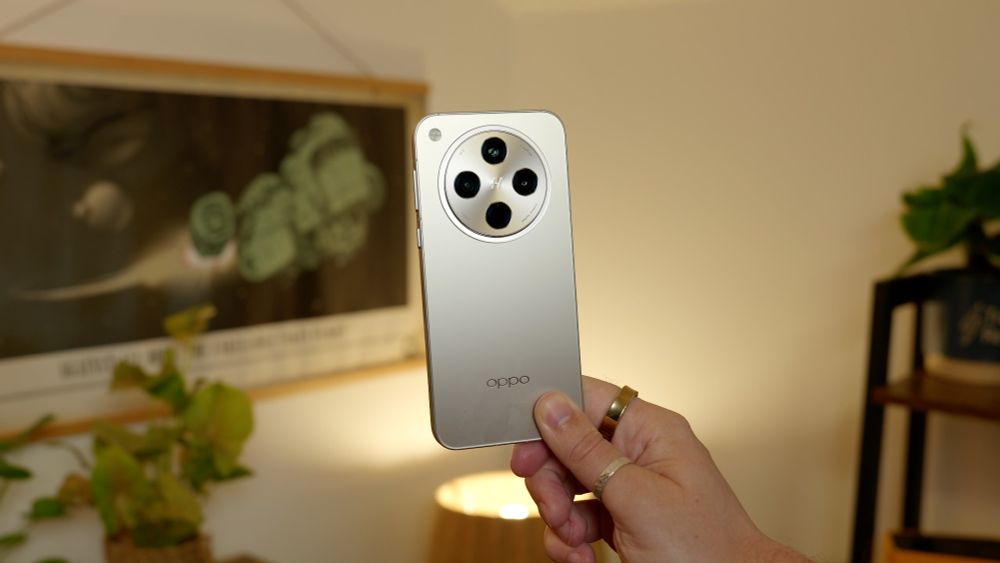 Smartphone being held centrally in a wide shot, blurred houseplants and poster in the background. Big round camera housing in the centre. 