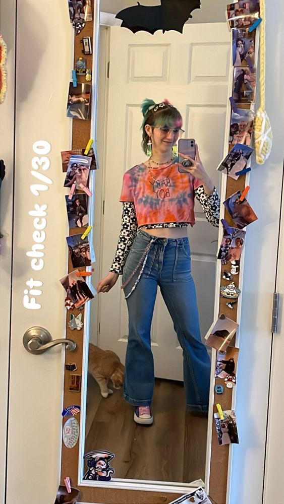 Jonesy poses in a full length mirror with text saying "fit check 1/30" on the side. Fae is wearing bell bottom jeans, a black and white long sleeve patterned crop top under a pink tie-dye crop top. Their short split dye pink and blue hair is half up in two pigtails with star clips. They are also wearing ufo earrings, star glasses, two colorful belt chains, and rainbow converse. 