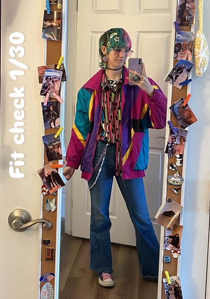 Jonesy, a thin AFAB person with split dye pink and blue hair poses in a mirror. The words "fit check 1/30" are on the side of the image. Fae is wearing flare jeans, and multicolored patterned shirt and magenta, teal, purple, and yellow windbreaker. They have large patterned statement earrings, star hair clips, and two toned pink and blue star glasses. 