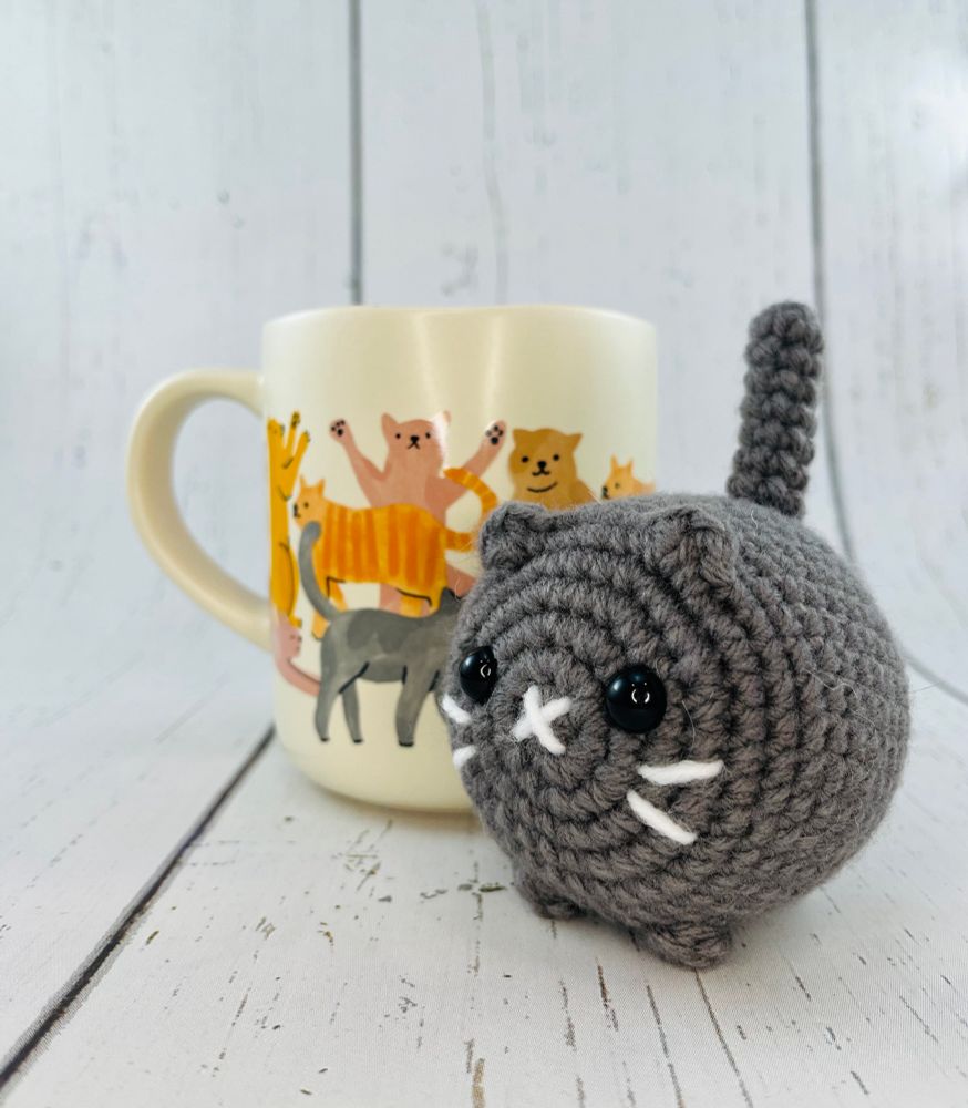 Mini crochet cat beside a mug. The mini cat is shorter in height, length and width than the mug. The cat can fit inside of the mug.