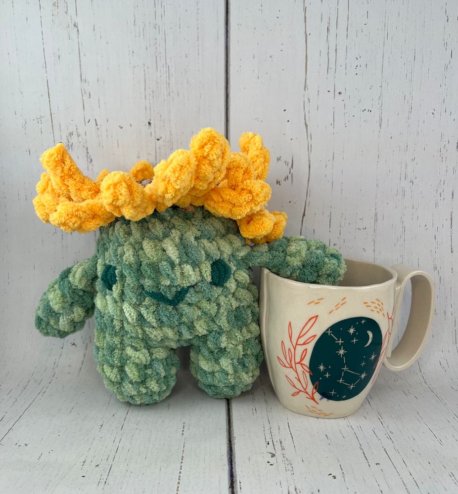 A crochet squatch is next to a mug. A squatch is a cute creature with arms and legs. It is slightly taller and wider than the mug. It has a sunflower on sewed its head.