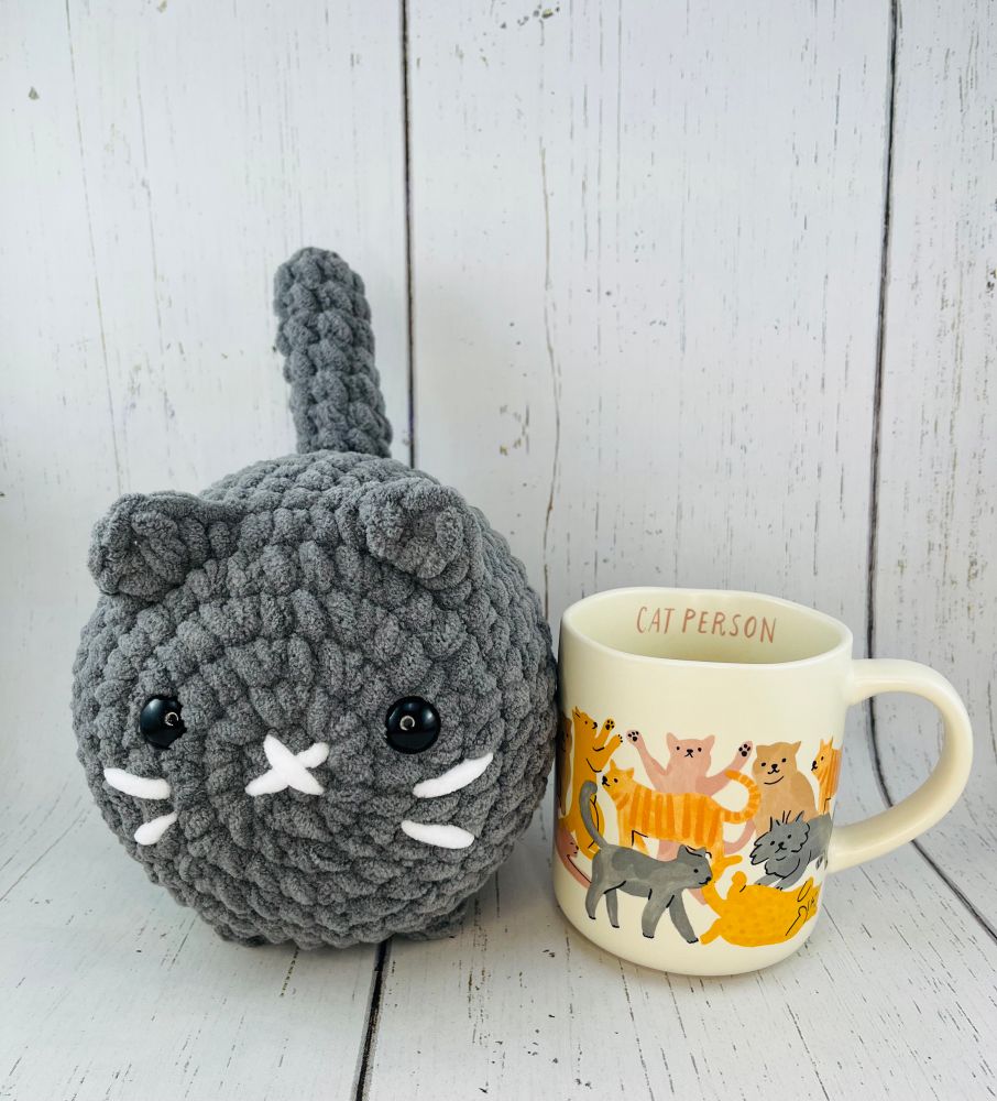 Picture of big crochet cat beside a coffee mug. The crochet cat is taller, longer and wider than the mug. 