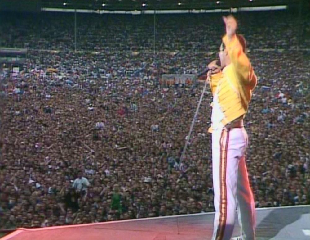 Queen live at Wembley in 1986. My first gig, I was 15.  Camera catches Freddie Mercury from behind, mic held in front of his mouth, left fist raised, as he conducts the crowd.