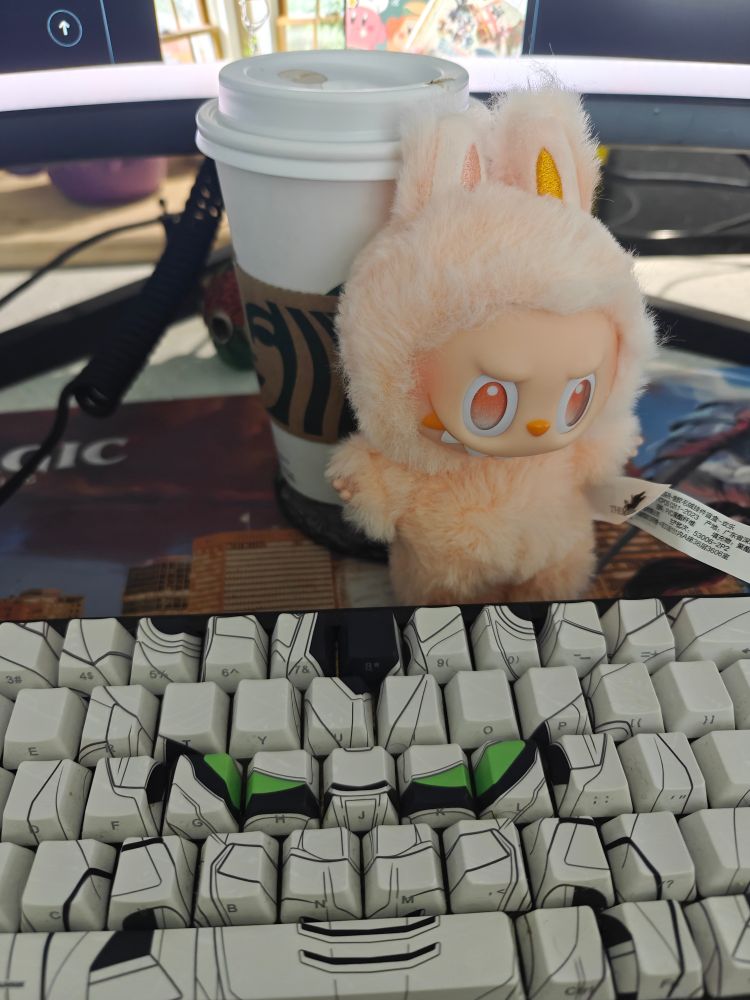 a happiness Big Into Energy labubu posed against a Starbucks cup in front of a Higround Gundam Wing keyboard, which is on top of a MTG pro tour dominaria mat 