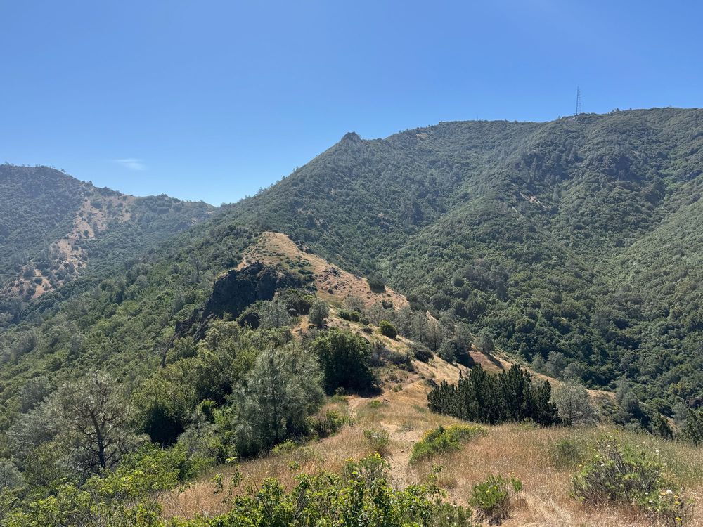 A rocky dirt trail leads the eye higher through a series of hills covered in scrubby bushes under a brilliant blue sky.
