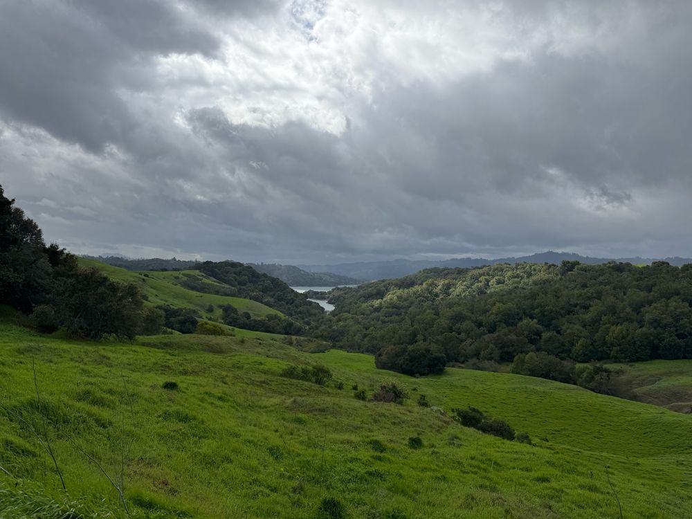 Rolling green hills dotted with scrubby oaks in the foreground lead to a reservoir in the background. The sky is full of clouds, alternatingly dense and wispy, ranging from dark blue-gray to bright white. 