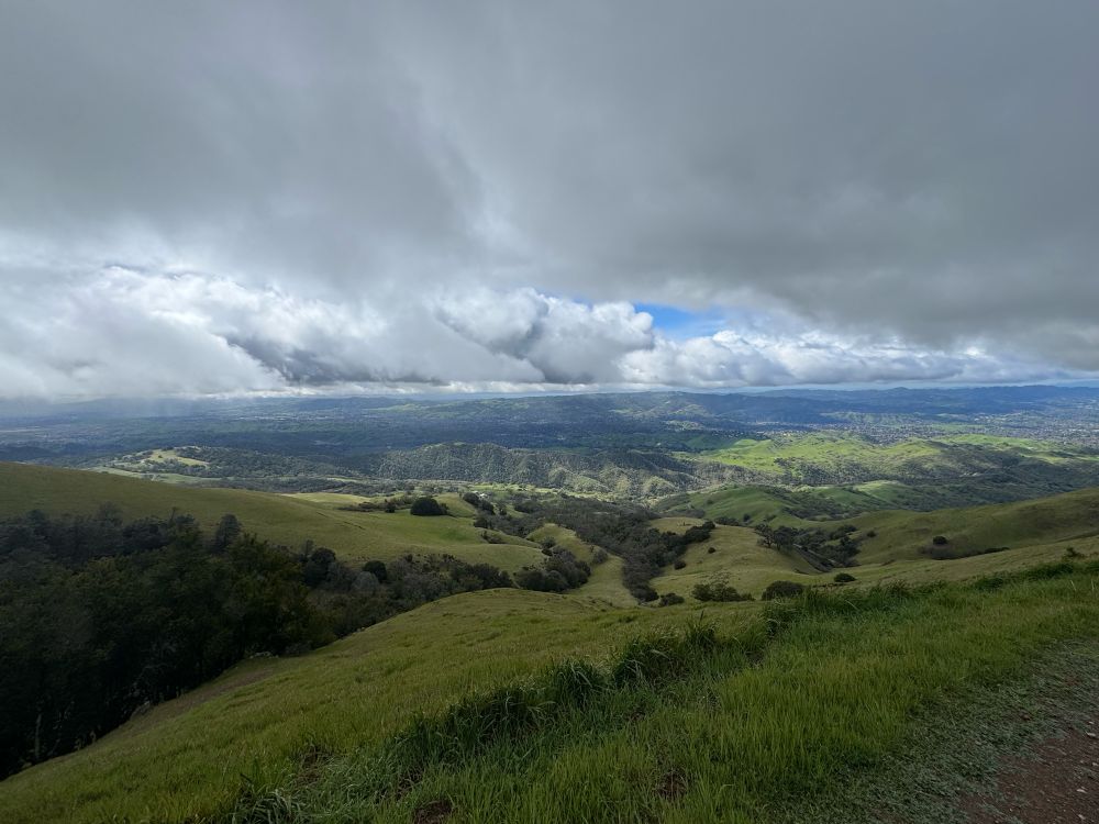 Above, thick white and gray clouds part to reveal a patch of striking blue sky. Below, bright green hills fold over each other.