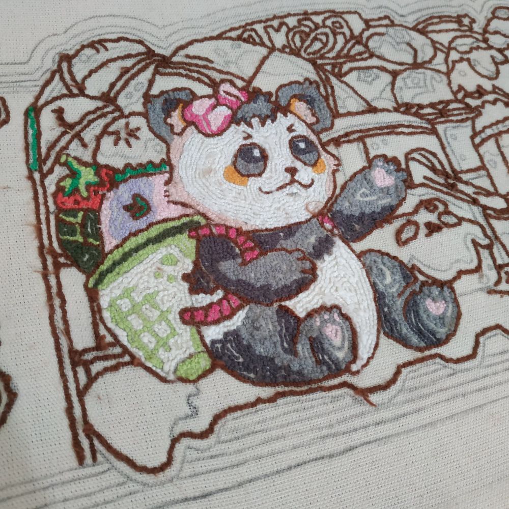 Parte traseira de um tapete bordado em ponto russo (punch needle) em andamento. Um panda com um laço rosa na cabeça e blush nas bochechas em laranja. Seu pelo tem dois tons de cinza, além do rosto branco e sombras em cor de pêssego. Sua mochila de bambu está cheia de alimentos diversos. O resto da imagem ainda continua em progresso, mostrando apenas os contornos bordados em marrom.