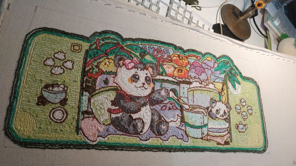 Parte traseira de um tapete bordado em ponto russo (punch needle) com 99% do bordado concluído. A imagem geral representa o formato e estética de um nintendo switch. O cenário principal é bastante colorido com dois pandas, adulto e filhote, dispostos ao na frene de frutas, vasilhames com bolinhos de massa (dumplings), bolos, cremes e um copo de suco. Os desenhos nas extremidades do tapete representam os botões do console mas em formatos de comida. O tapete é tem vários tons de verde, sendo a cor que mais prevalece.