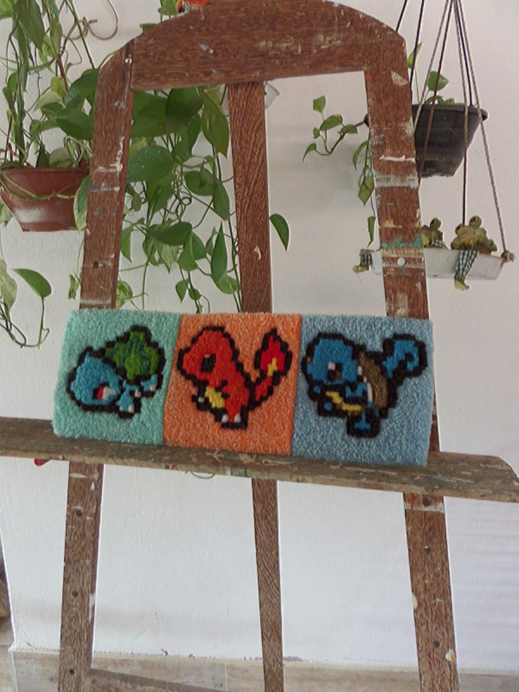 Fotografia de um tapete de teclado retangular, feito em ponto russo (punch needle), apoiado em um cavalete de madeira manchado de tinta, na frente de vasos de plantas. O tapete é colorido e tem o design em pixel art de três pokémons: bulbasaur, charizard e squirtle.