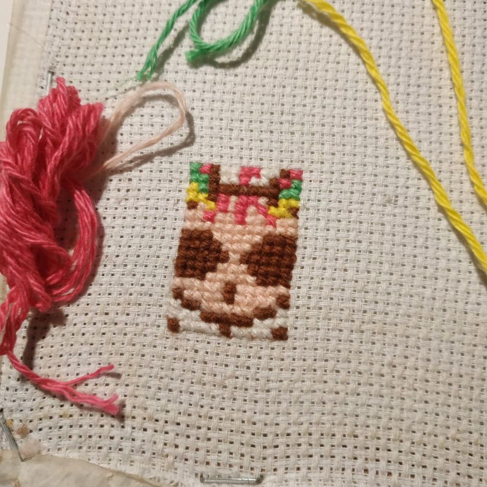 Fotografia com close-up de bordado retangular em ponto cruz da parte central focada de um rosto cartoonizado bastante sorridente, com olhos escuros bem grandes e fios de cabelo rosa. Há alguns fios de lã soltos para compor a fotografia ao redor do bordado nas cores rosa, verde e amarelo.