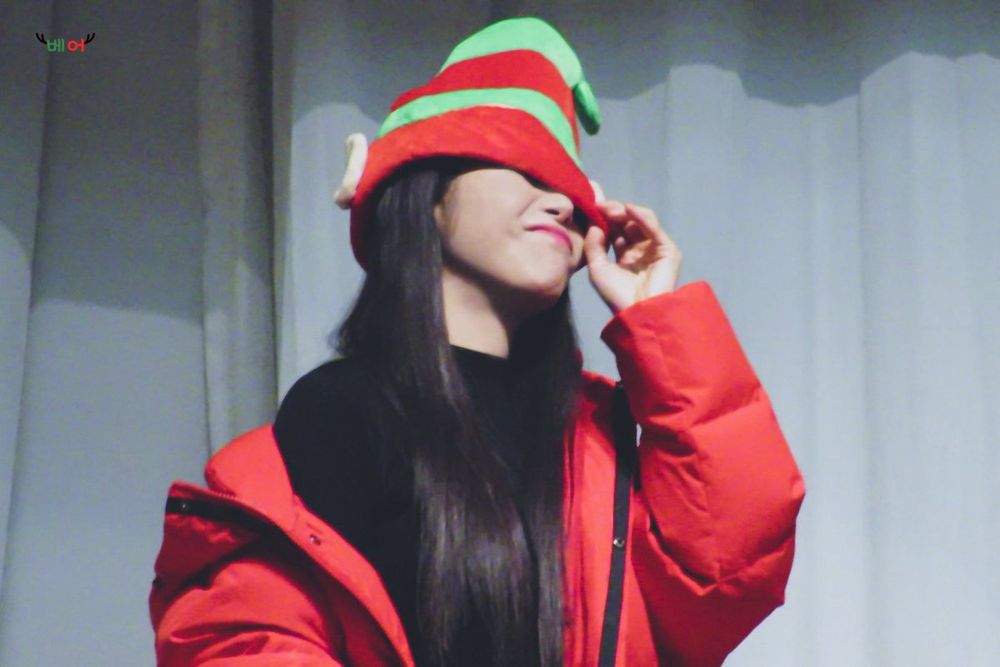 Solar está usando um casaco vermelho com um chapéu de elfo verde e vermelho. O chapéu cobre parte do rosto dela, e ela sorri levemente enquanto ajusta o chapéu com uma mão.