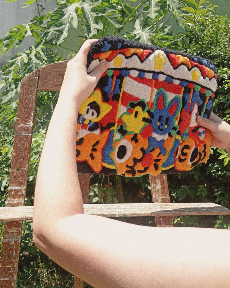 A fotografia retrata o momento em que eu, Giulia Congê, estou colocando um tapete colorido em um cavalete marrom manchado de tinta por uso, em um quintal bem verde, mas só meus braços estão a mostra. O tapete foi feito em ponto russo (punch needle), com cores em tons saturados em um fundo preto. Os personagens ilustrados são originais, feitos pelo artista Toriko. A ilustração mostra os personagens acomodados e se divertindo em um carrossel, em cima de assentos com formatos de grandes peixes laranjas com olhos grandes, redondos e bem abertos. 