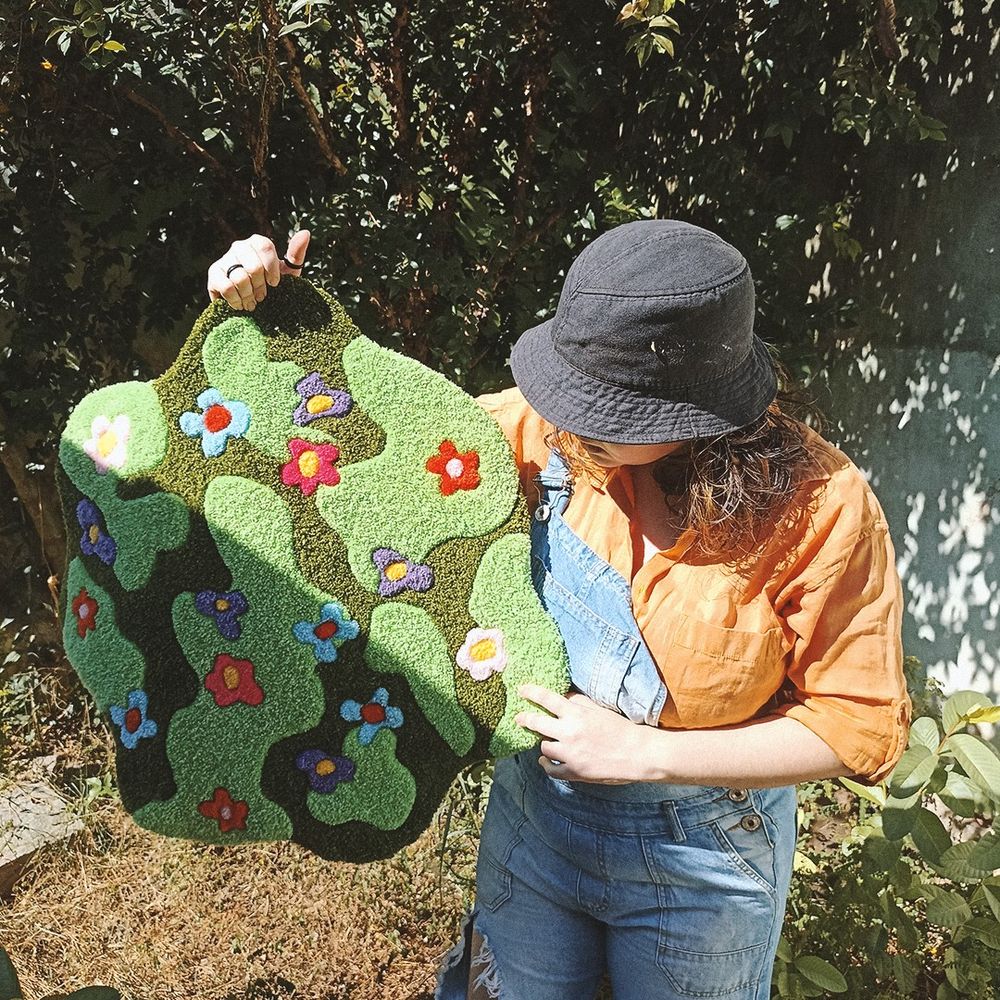gegê congê está vestindo uma camisa laranja e um macacão jeans e segura um grande tapete de punch needle com um padrão orgânico e curvilíneo, imitando um jardim. o tapete é predominantemente verde com flores coloridas em tons de roxo, rosa, laranja e azul.