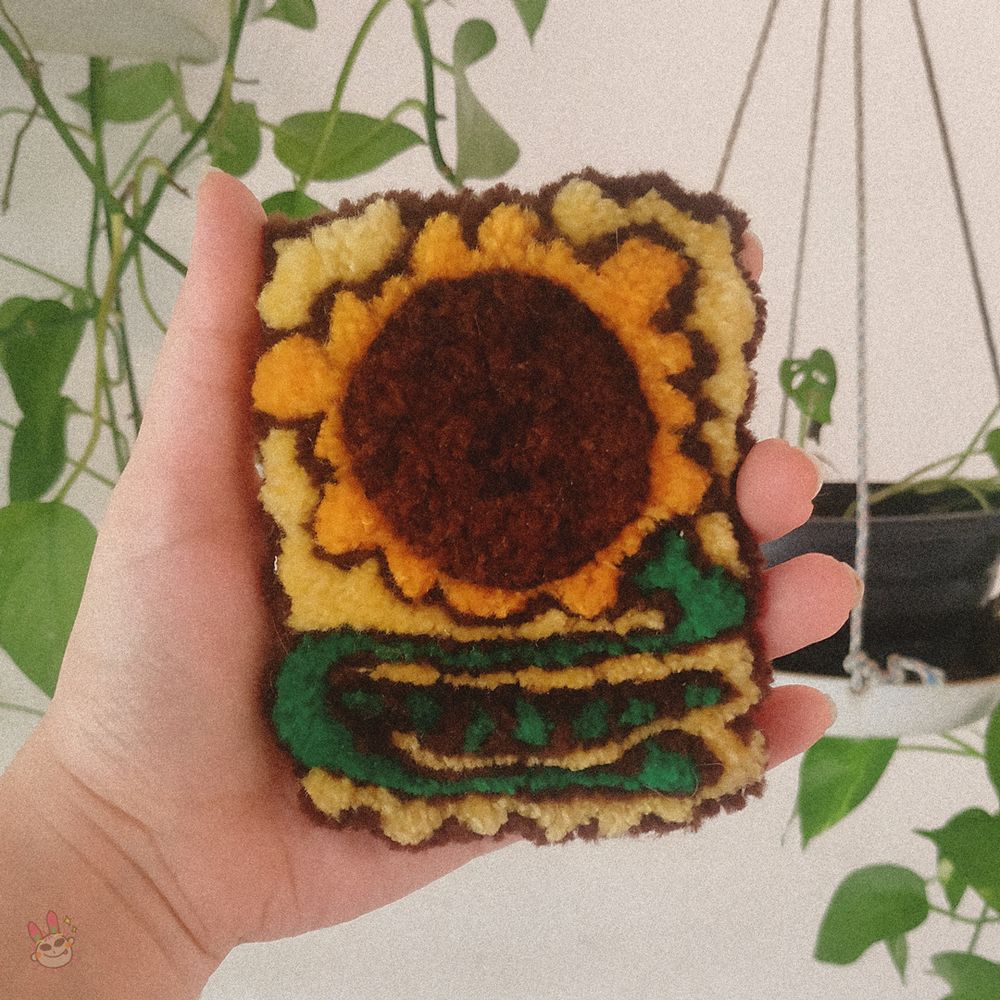 Fotografia de um broche de lã feito em ponto russo (punch needle) retangular de um girassol estilizado em um fundo amarelo com caule verde curvado, miolo marrom redondo bem grande e pétalas amarelas. Uma mão o segura na frente de algumas plantas e uma parede branca.