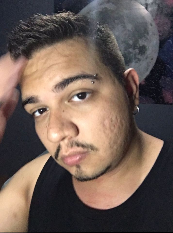 Homem branco com regata preta, piercing na sobrancelha, e no fundo uma pintura de uma lua cheia