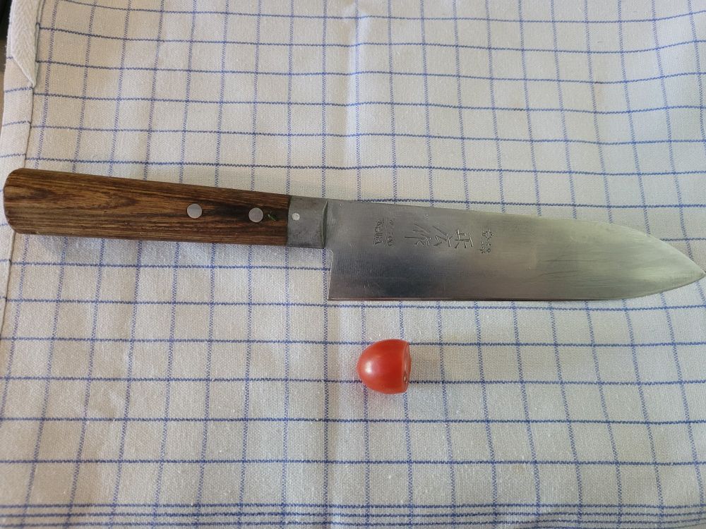 En innmari fin og godt brukt japansk kokkekniv av merket Masahiro på et kjøkjenhåndkle med en halv cherrytomat under.