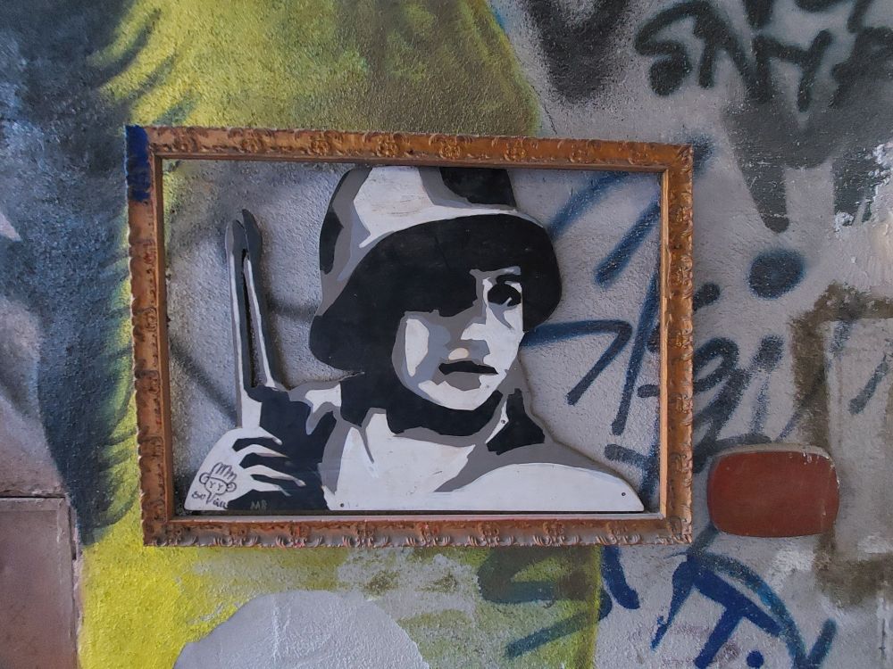 A B & W & grey stencil image of a closeup of a woman wearing an army helmet and carrying a rifle. Underneath that image is graffiti that's black, blue, and yellow.