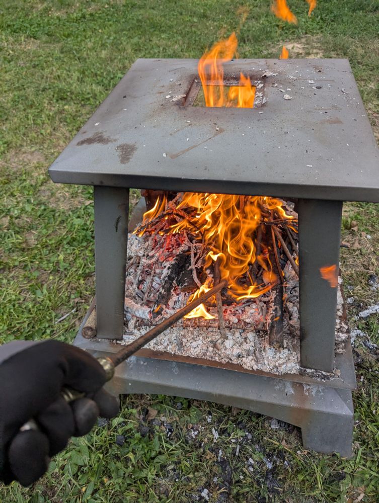 fire pit with a fire going, a hand with a black glove is stoking it with a metal rod