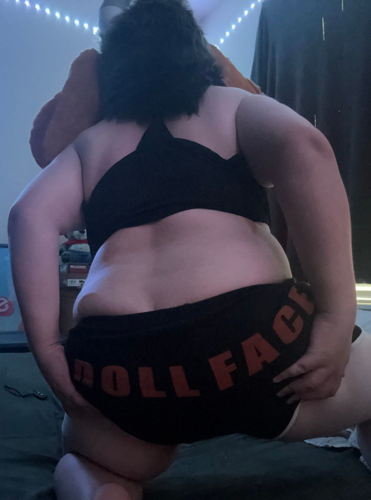 A photo of Daisy IRL, on her knees with her back to the camera, wearing only  a sleeveless black top, and booty shorts with the word “DOLL FACE” written on the butt area. Her hands are supporting her cheeks, as she bends over slightly on her bed.