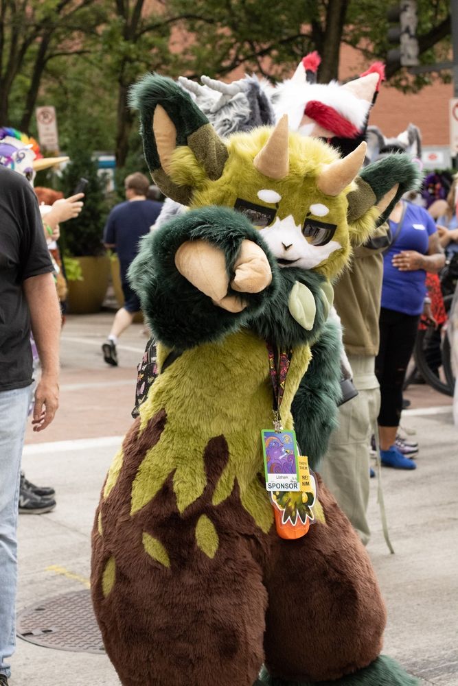 Image of a fursuiter (me) pointing at the photographer

The fursuit is a (from bottom to top) Brown, green, do deep forest green, goat like fae creature. It's body is leaf patterns. The head has large, wide eara similar to a deer, and small sharp horns like a domestic goat.

The fursuit is in a kemono style, so large domed eyes, little mouth and nose.
