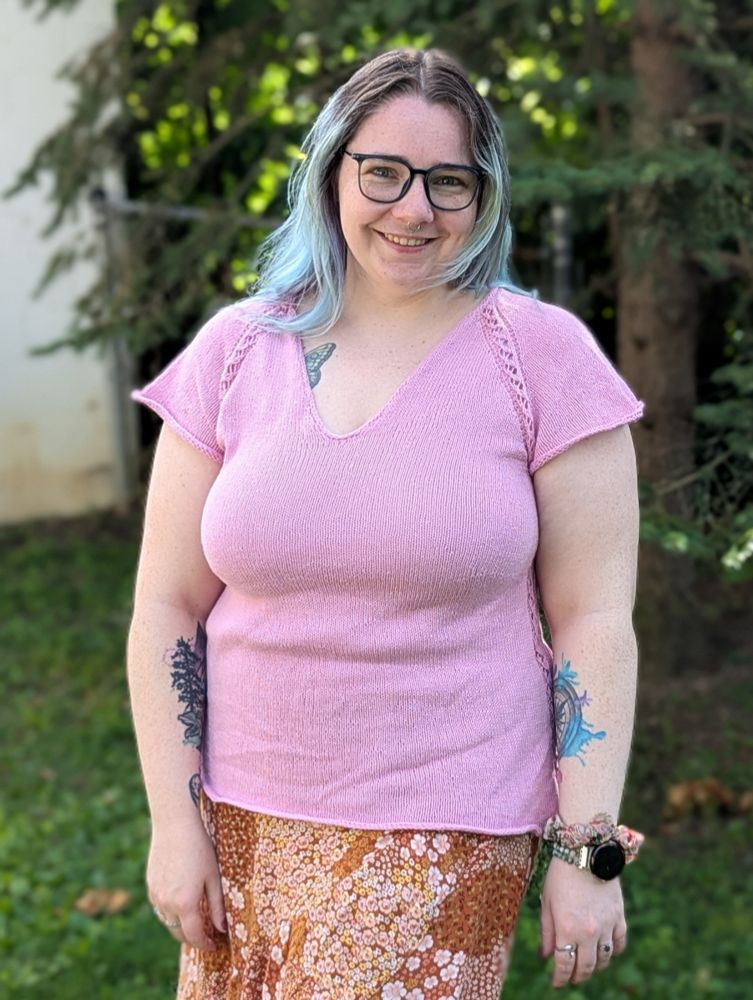 A white woman with blue and purple hair and blue glasses stands in front of deep green evergreen trees. She wears a light pink knitted v-neck tee shirt with lace details along the raglan and a brown and pink floral floor-length skirt.