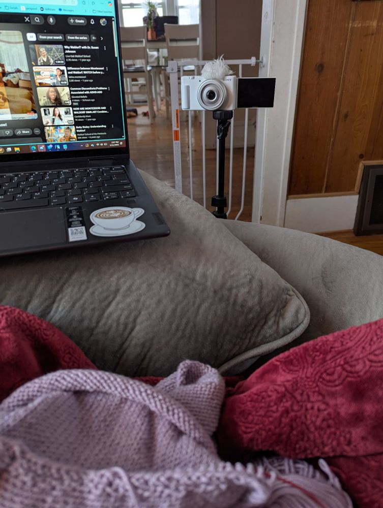 This is a point-of-view photo of a grey couch with a fuzzy red blanket covering the lap of the photographer. A black laptop with a coffee sticker can be seen to the left. On the person's lap sits a lilac knitting project, the start of a sweater. Straight ahead a white camera on a tripod can be seen. 