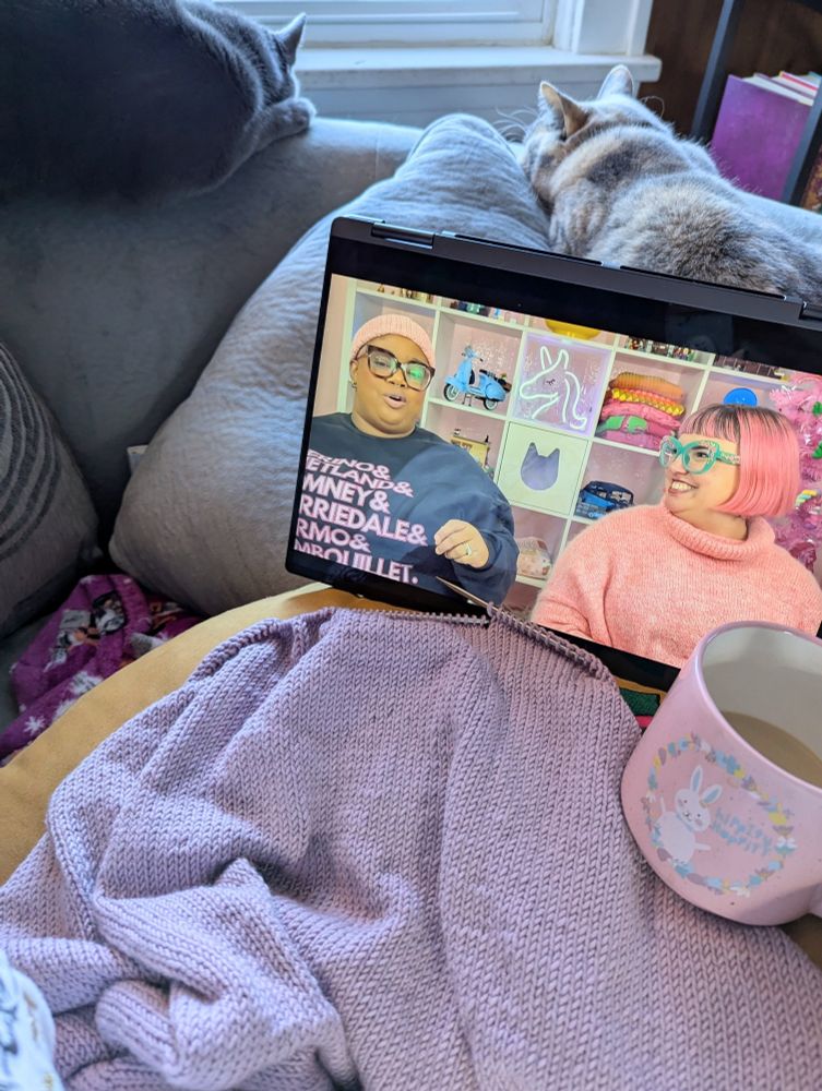 Kaity sits on a grey couch. A purple knitted sweater rests on her lap next to a pink coffee cup with a white bunny that says "hippity hoppity". On a tablet screen we see a still of a podcast where Shayla- a Black woman with black glasses, a sweatshirt that lists sheep breeds, and a pink knitted hat- and Heather- a white woman with neon pink hair, a matching sweater, and teal glasses - sit in front of a vibrantly pink background to discuss their knitting projects. Behind the screen, we see two cats lounging and looking out the window.