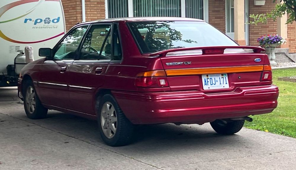 Left Rear 3/4 view of a red, mid 90’s Ford escort LX 5 door parked in a driveway 