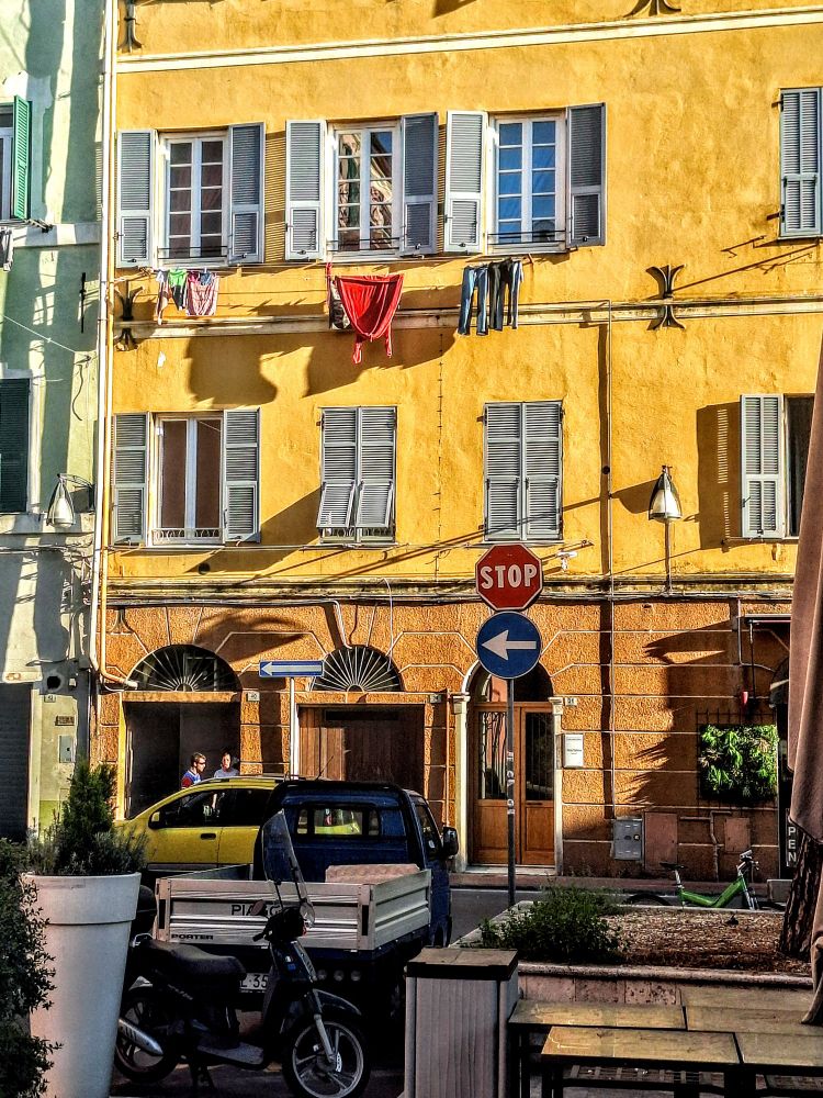 Hell beschienene Häuserfront. Gelbe Hauswand, Fenster mit grauen Läden. Es hängt Wäsche an der Hauswand. Weiter unten hat es Verkehrszeichen. Unten auf def Strasse verschiedene Verkehesmittel, Autos, Ladepiaggos, Mopeds.