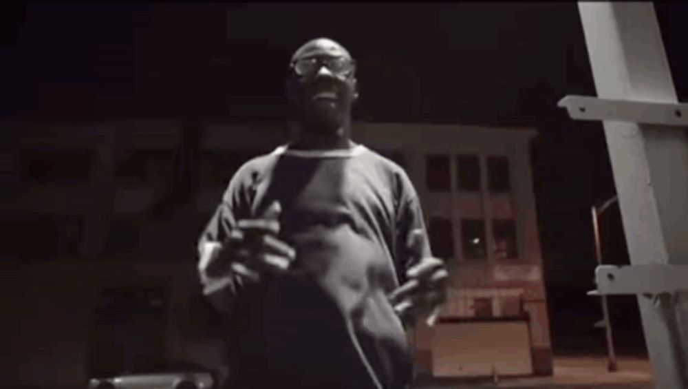 a man in a red t-shirt is standing in front of a brick building with his arms outstretched .