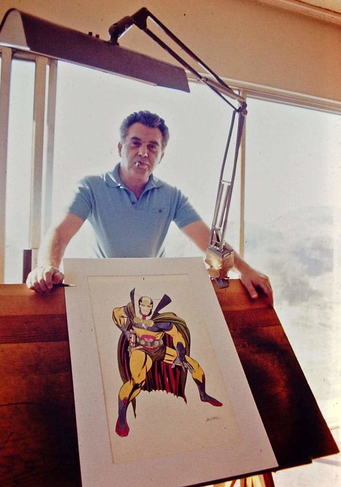 Jack Kirby standing behind his drawing table. He has his hands on the desk, and there is a paper with a colored drawing of Mister Miracle on the table.