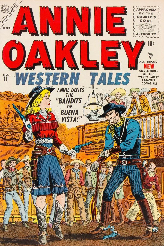 Annie Oakley Western Tales #11 (1956). On the cover, Annie is in a saloon and has just shot the gun out of the hand of her opponent. He rubs his wound and scowls at her. There is a large crowd of cowboys in the background watching the scene.