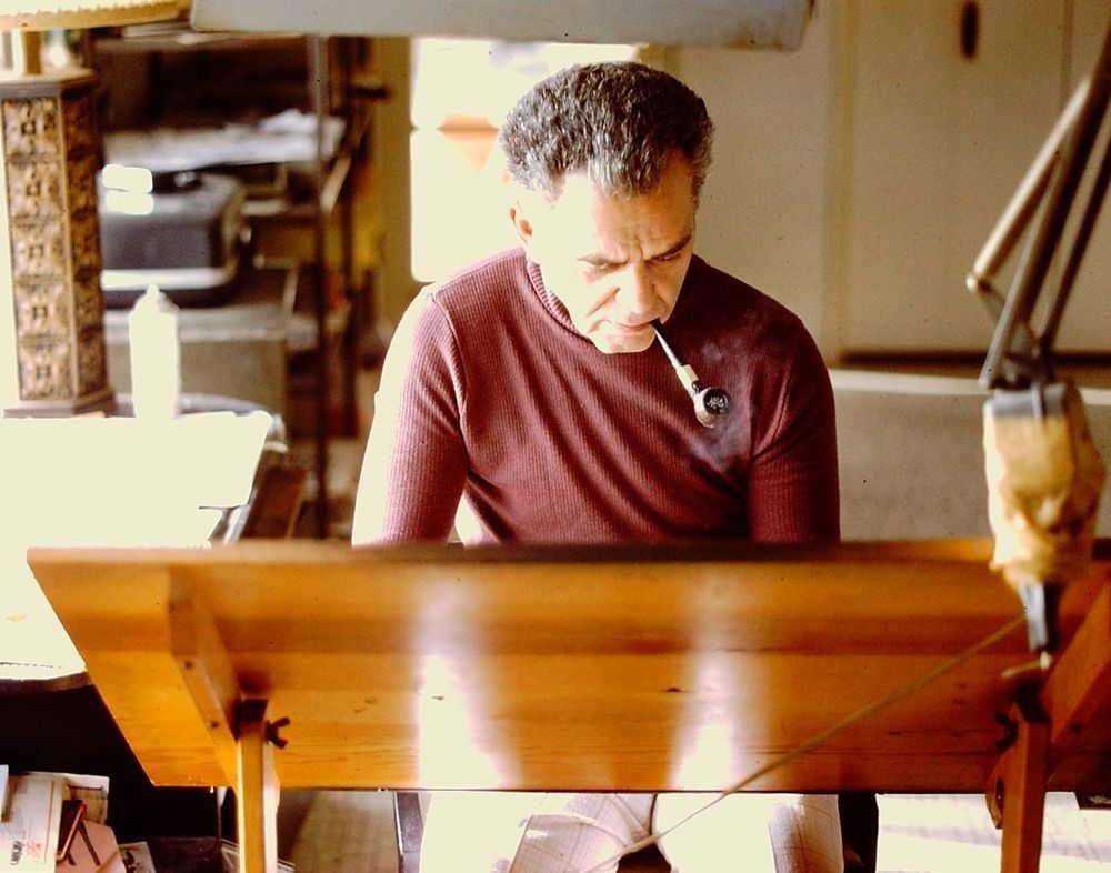 Jack Kirby at his drawing table with a pipe in his mouth.