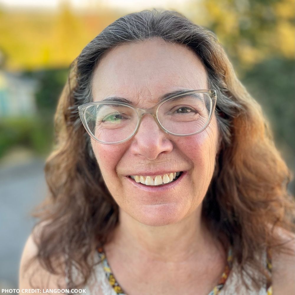 A photograph of the late poet Martha Silano. She is a middle-aged woman with long, wavy brown-grey hair and clear-rimmed glasses. She is photographed outside, with leaves/trees behind her.