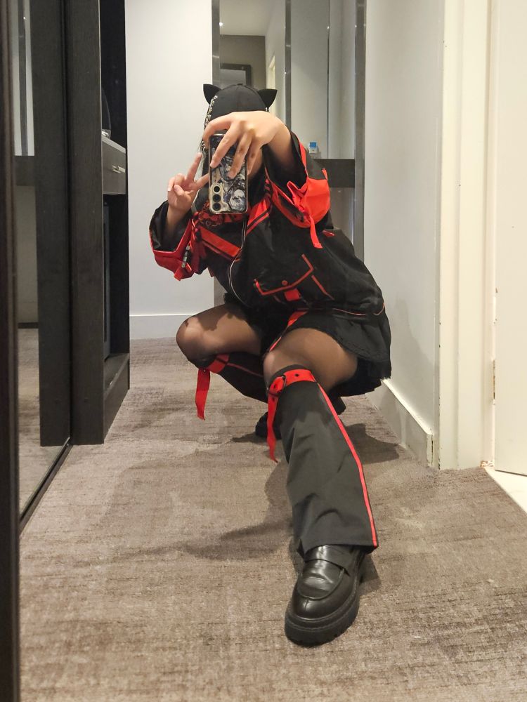 Me, taking a mirror selfie of my outfit. I am wearing a red and black jacket, a black skirt, matching leg warmers and loafers. I am also wearing a black cap with cat ears. I am squatting down near the floor with my left leg out for balance. My phone is held in a way that obscures my face, and am posing with a peace sign.