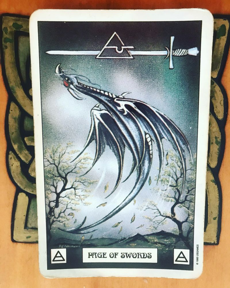 A blue/gray dragon flying in the air looking to be doing a somersault or backflip. There are 2 small trees and a mountain range in the background. A single sword floats horizontally at the top of the card