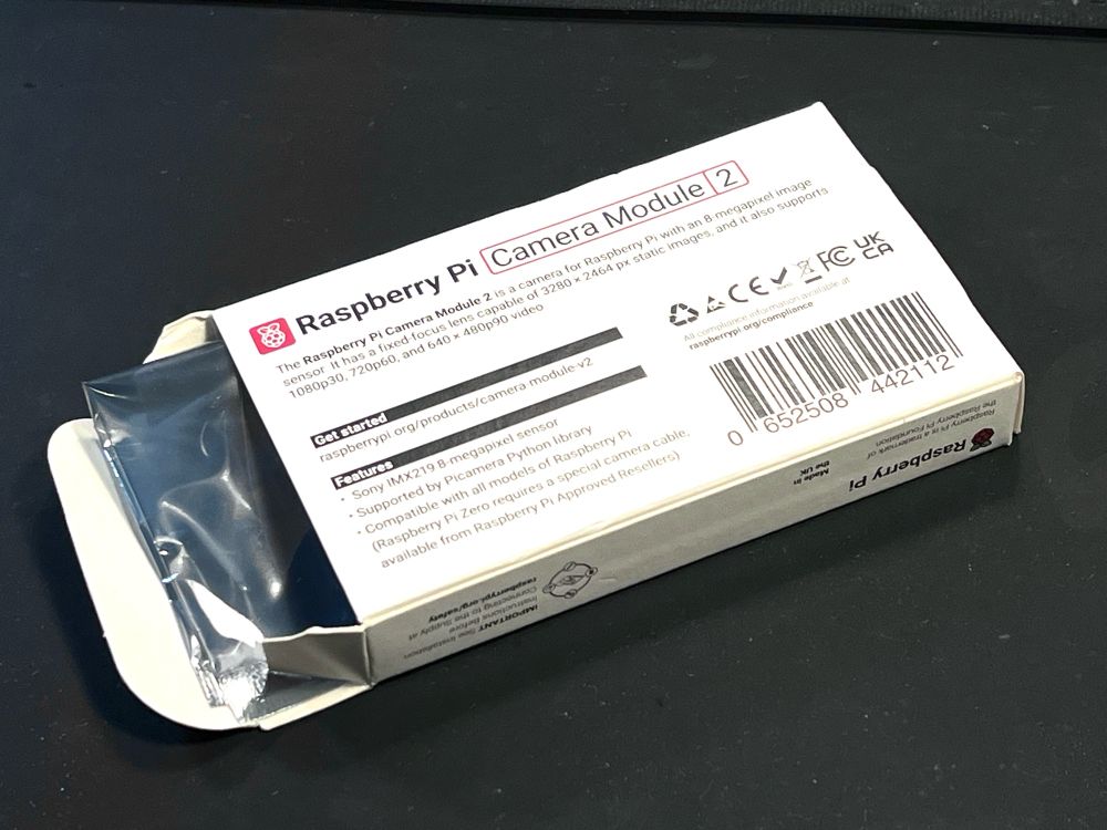 A small white rectangular paperboard box opened on a black table with a silver plastic packet visible in the opening. The box says Raspberry Pi Camera Module 2 in a sans serif font with small print and a bar code that evokes a pharmaceutical box. 