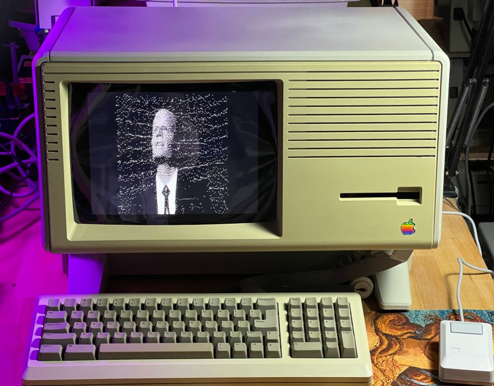 An Apple Lisa with Mac keyboard and mouse in front with Max Headroom on the screen in black and white dithering.