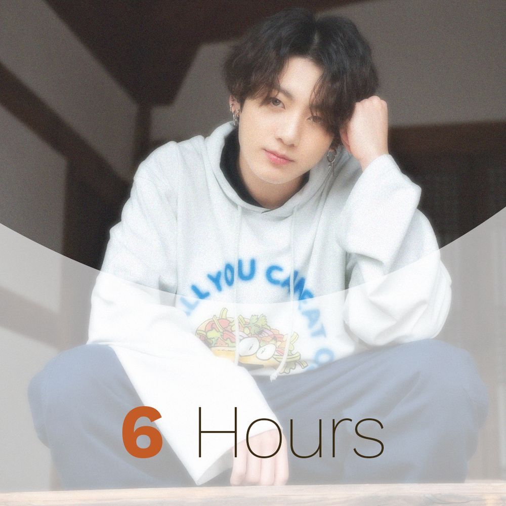 JK with dark wavy hair, wearing blue pants and a white hoodie with an animated salad and the words All You Can Eat on it. He is crouching down staring directly at the camera resting his head against one hand. The photo has a soft filter applied and says the words 