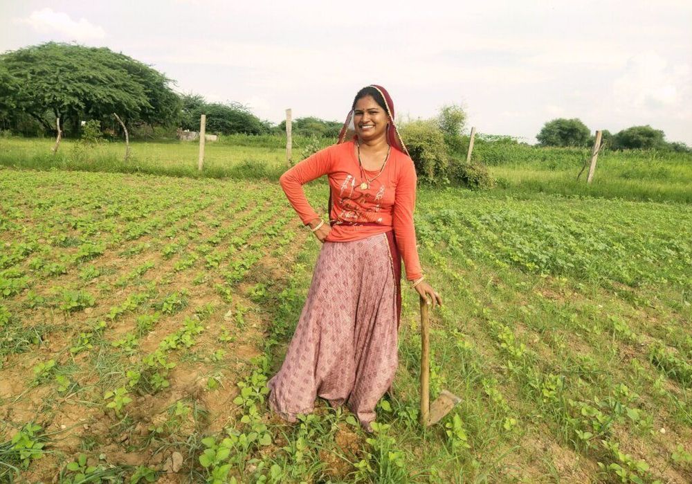 An Indian woman standing in a field of green farmland. She is looking at the camera smiling with one hand on her hip and the other resting on the hoe leaning on the ground.
