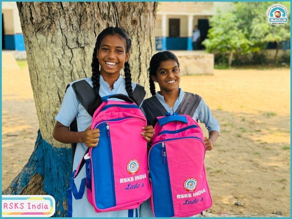 Two Indian girls standing in front of a large tree in what appears to bee their school yard. They are dressed in school uniforms, wearing RSKS India donated backpacks, and are looking at the camera with smiles on their faces.
