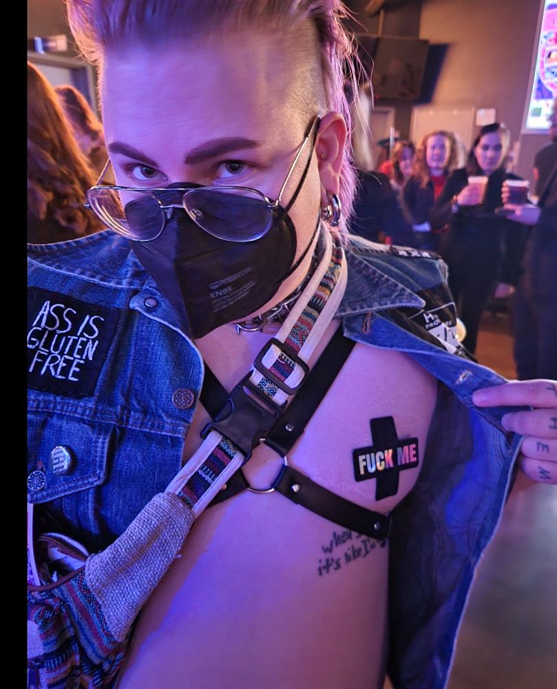 A white androgynous person with a purple mullet is holding open their jean vest to reveal a nipple pastie ghat reads "fuck me".
They have a black n95 mask, silver aviator framed glasses, big silver horseshoe earrings and have a leather harness across their bare chest.
A patch of their jacket is visible and reads "ass is gluten free"