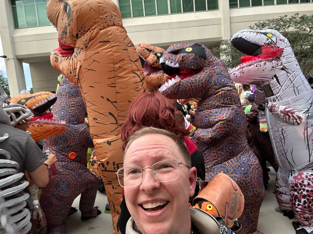 Goofy selfie of me (blonde, white 30-something man in rimless glasses) laughing while dancing on an outdoor patio surrounded by people in huge inflatable t-rex dinosaur suits of various patterns.