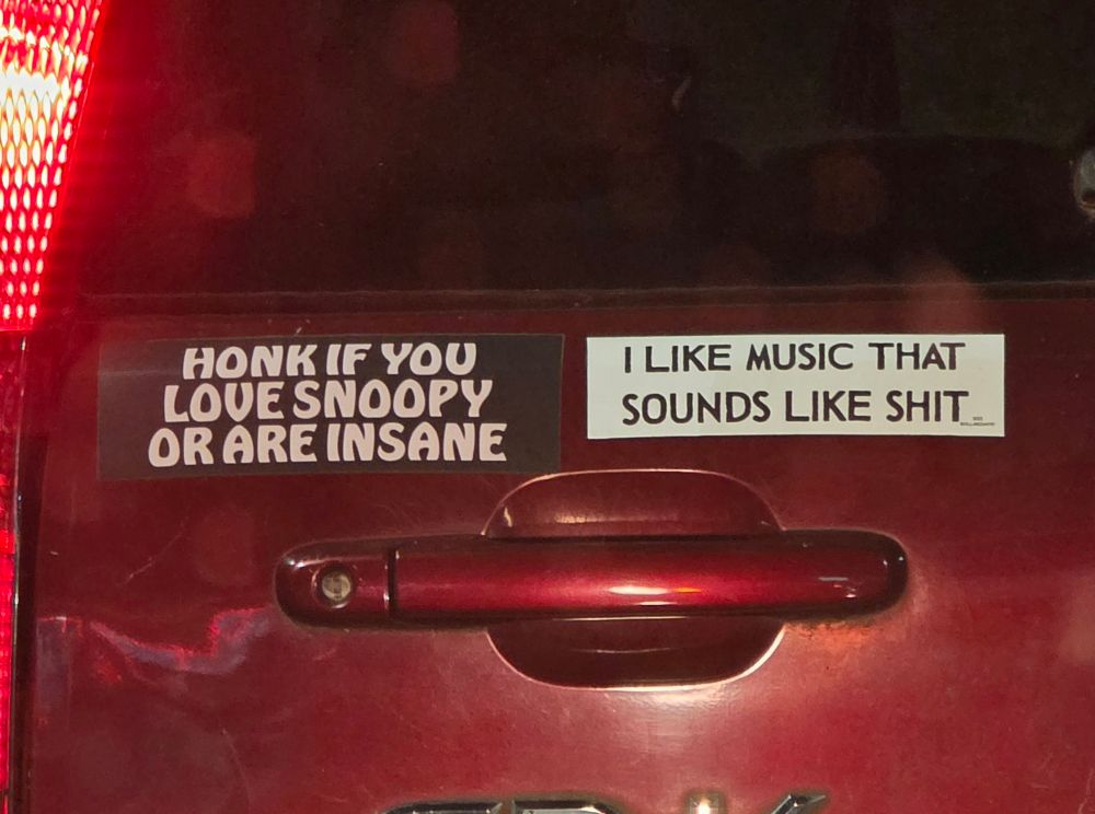 two bumper stickers on the back of a red Honda CR-V, one reads "HONK IF YOU LOVE SNOOPY OR ARE INSANE" and the other reads "I LIKE MUSIC THAT SOUNDS LIKE SHIT"