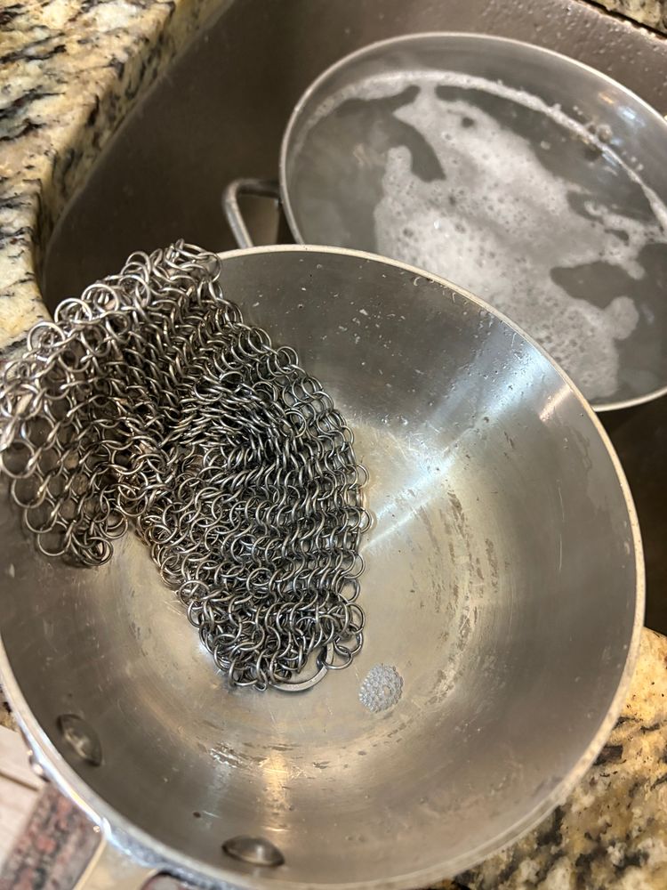 Chainmail laying inside of a clean pan. In the background there is a pot full of soapy water sitting in a sink